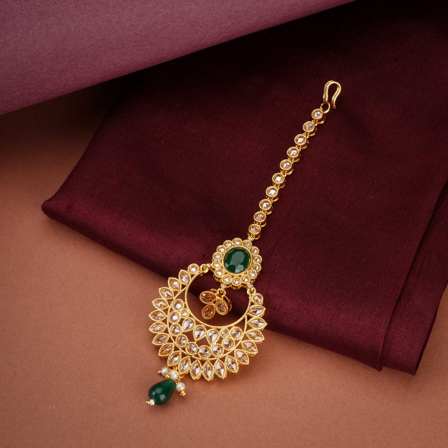 KUNDAN-STUDDED MAANG TIKA FEATURES INTRICATE WORK WITH A SINGLE GREEN PEARL AND A MATCHING STONE.