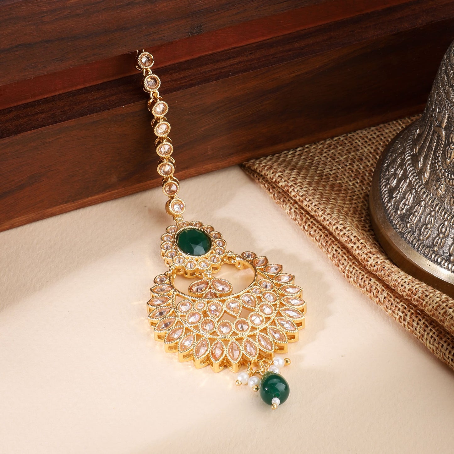 KUNDAN-STUDDED MAANG TIKA FEATURES INTRICATE WORK WITH A SINGLE GREEN PEARL AND A MATCHING STONE.